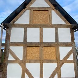 A partially constructed timber-framed building with boarded windows and a white facade. Completed using lime rendering. 