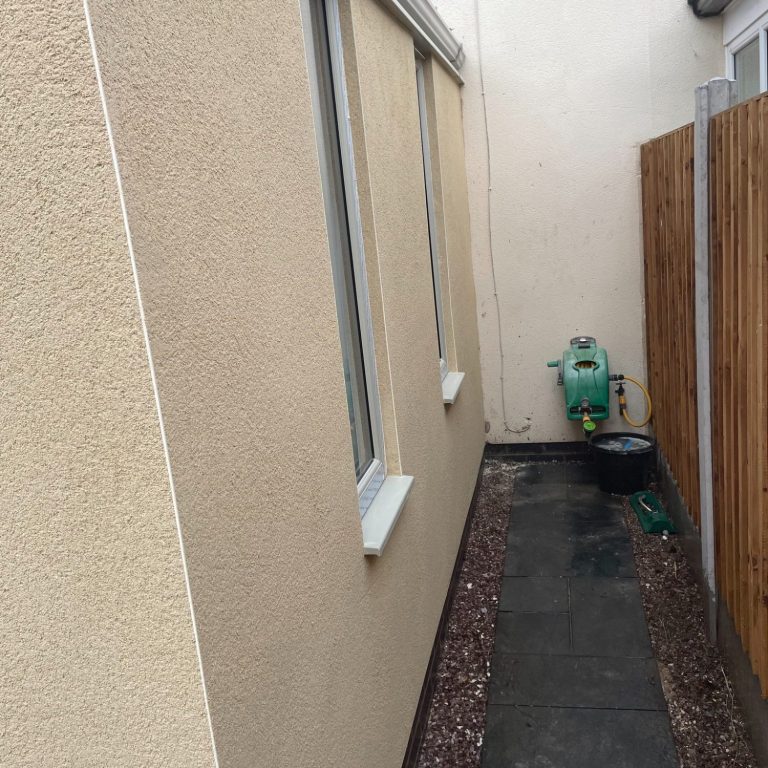 Narrow pathway between two buildings with a fence on one side. Monocouche rendering in ivory.